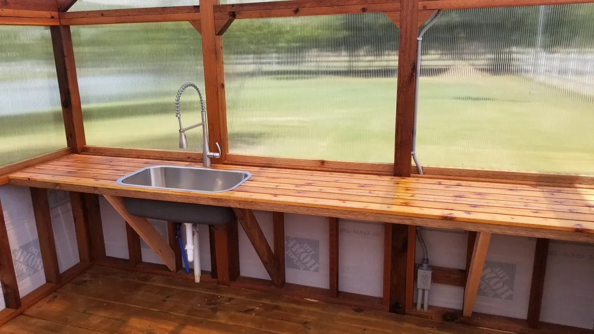 Cedar potting bench with an inset stainless sink and an electrical outlet below in a greenhouse